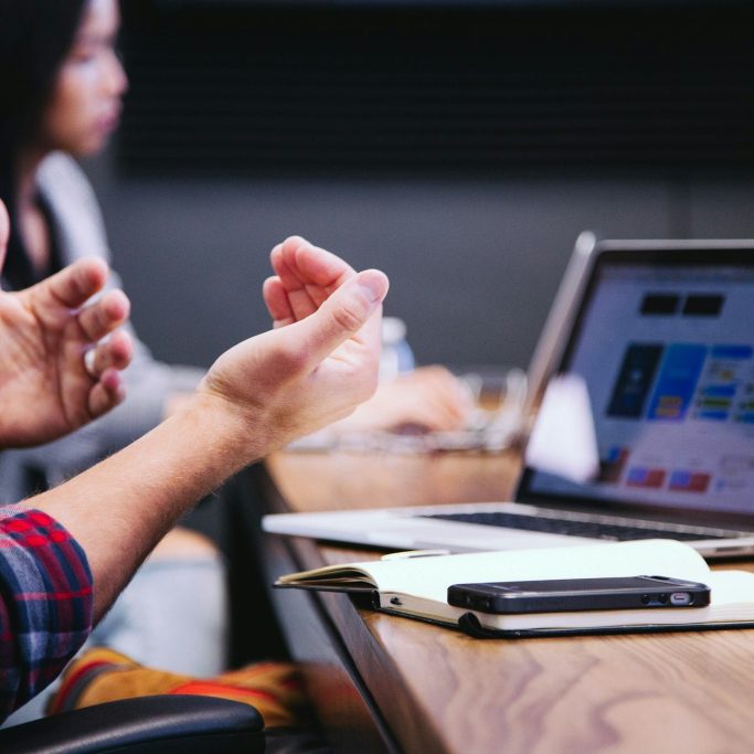 A person gesturing in a meeting with a laptop and notes on a table.