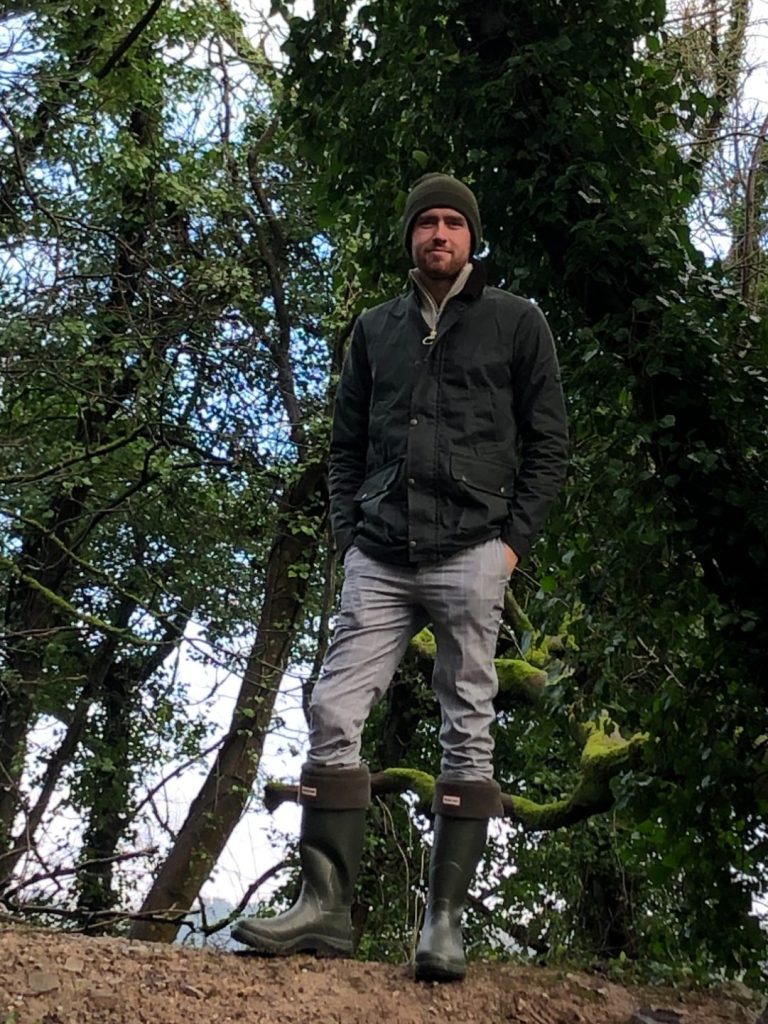 A man in boots and a beanie stands among trees in a wooded area.