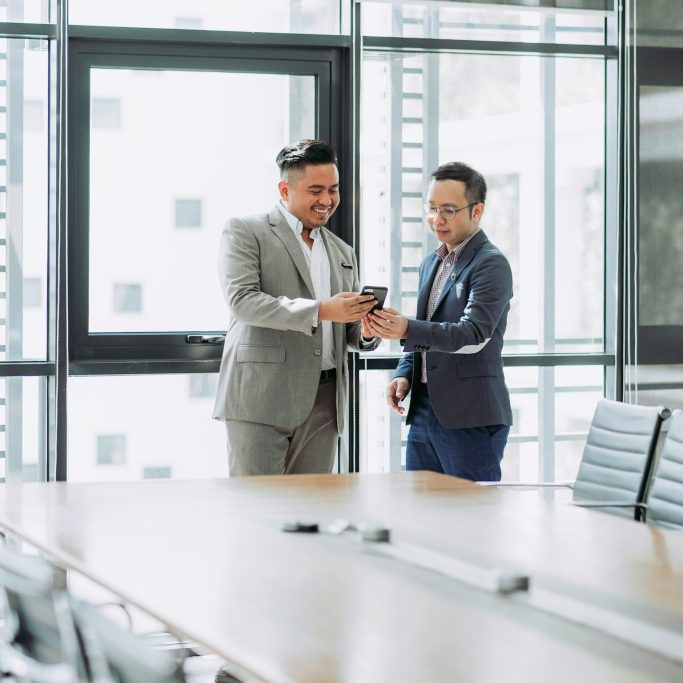 Two men in business attire collaborate while looking at a smartphone in a modern office.